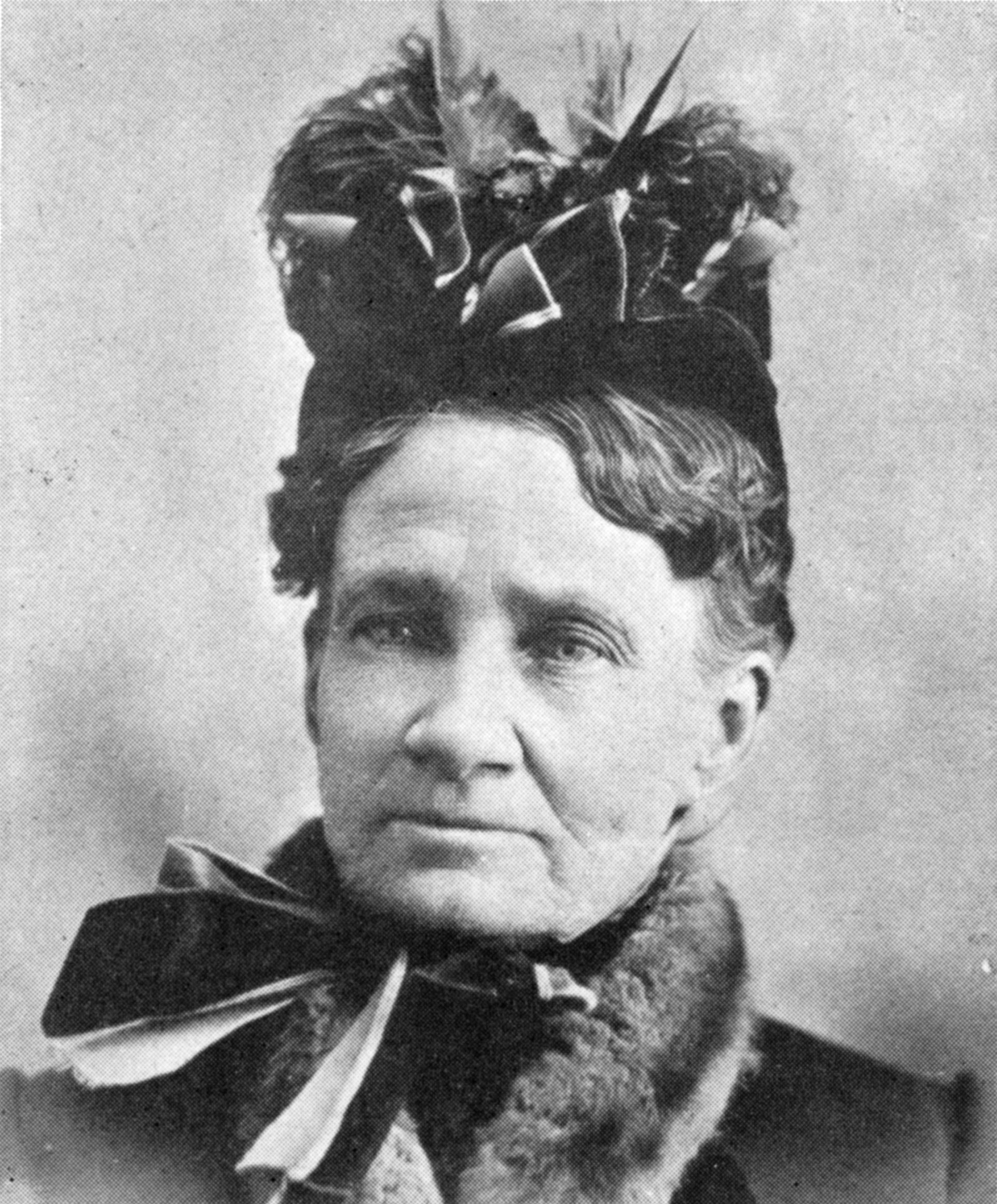 close up of woman's head, facing the camera, wearing a large hat and large bow at the neck, black and white.